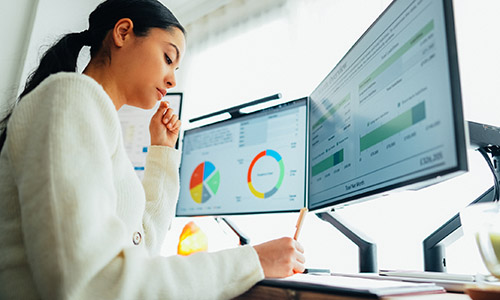 A woman looks at dual computer monitors with graphs and charts