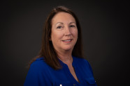 Woman with shoulder length dark hair parted on the side wearing a blue long sleeve shirt. 